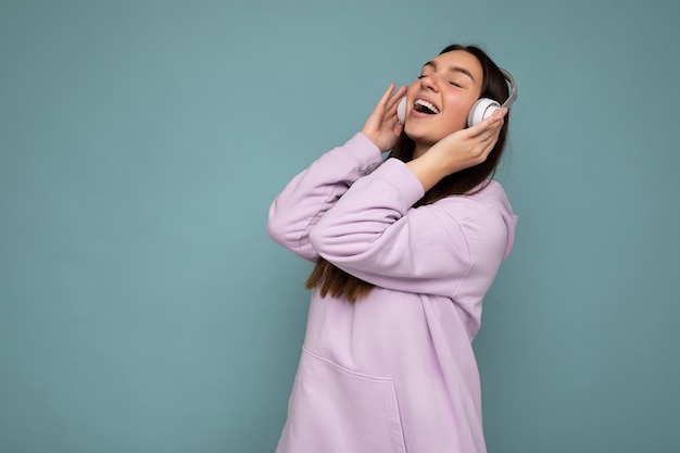 Ziemlich positiv lächelnde junge brünette Frau, die hellvioletten Kapuzenpulli trägt, isoliert über blauer Wandwand, die weiße Bluetooth-Kopfhörer trägt, die gute Musik hören und sich bewegend haben, Spaß zu haben. Speicherplatz kopieren