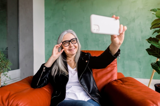 Ziemlich positive freudige 60-jährige Frau mit glattem grauem Haar, posierend auf ihrem Smartphone für Selfie-Foto, sitzend im roten Sessel vor grüner Wand im Studio