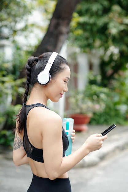 Ziemlich positive junge Frau mit Kopfhörern, die Musik auf ihrem Smartphone einschaltet, um sie während...