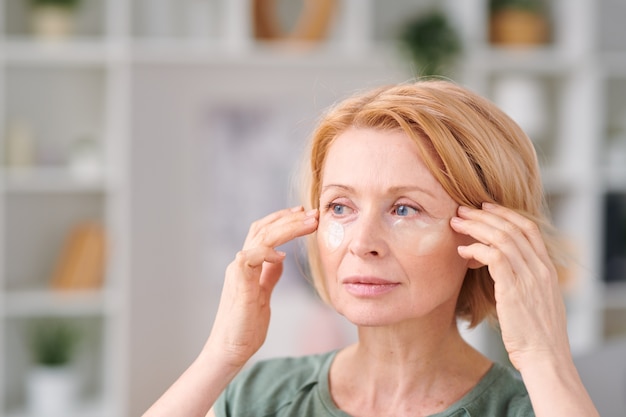 Ziemlich reife Frau mit kurzen blonden Haaren, die Unteraugenflecken setzen und sich während der Selbstisolation zu Hause um ihr Gesicht kümmern