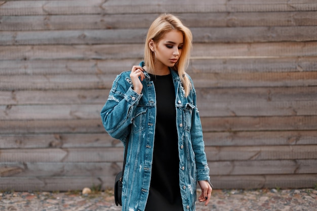 Foto ziemlich schöne junge frau blondine in einem schwarzen kleid in einer langen jeansjacke mit einer modischen schwarzen lederhandtasche steht draußen in der nähe einer vintage holzwand