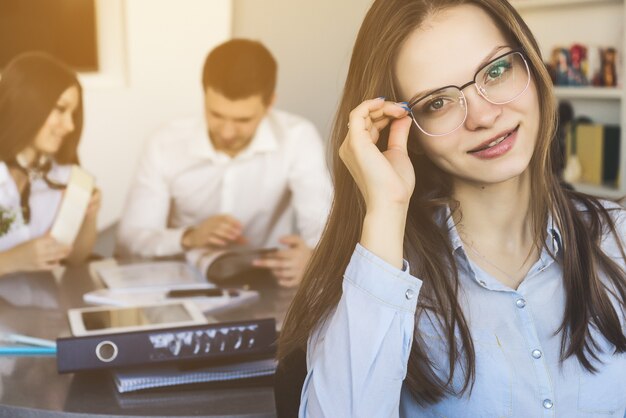 Ziemlich selbstbewusste Bürofrau mit Brille, die über den Hintergrund mit sprechenden Kollegen lächelt. Lehrerin hautnah in der Klasse