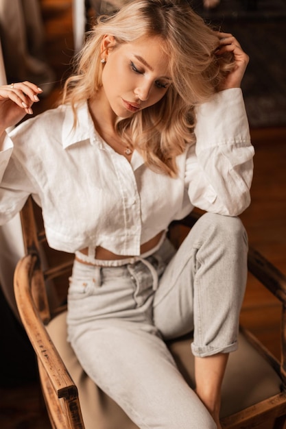 Ziemlich sexy blonde Frau in weißer Modekleidung mit Hemd und Jeans sitzt drinnen auf einem Vintage-Holzstuhl