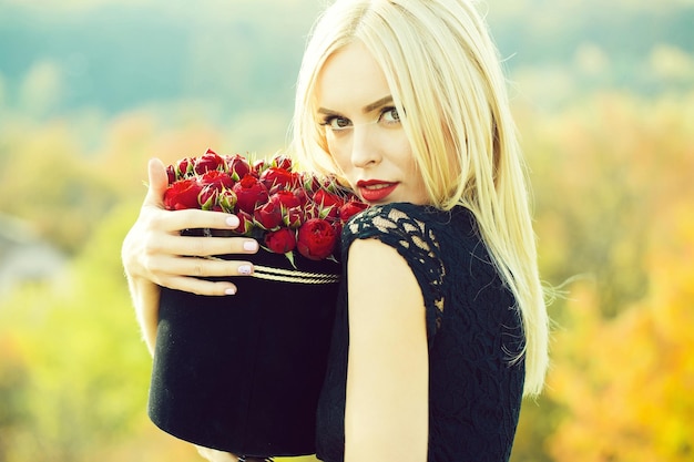 Ziemlich süßes Mädchen oder schöne blonde Frau in sexy schwarzem Kleid mit roten Rosenblüten im Feld im Freien auf natürlichem Hintergrund