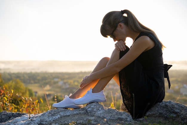 Ziemlich traurige Frau im schwarzen kurzen Sommerkleid, die auf einem Felsen sitzt und bei Sonnenuntergang im Freien denkt. Modische Frau, die an einem warmen Abend in der Natur nachdenkt.