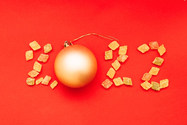Ziffer 2022 aus bunten Süßigkeiten und goldener Weihnachtskugel auf rotem Grund