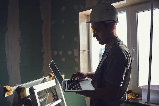 Zimmer reparieren. Baumeister mit Helm und Brille steht im Profil mit einem Laptop in der Hand auf dem Hintergrund der Baustelle