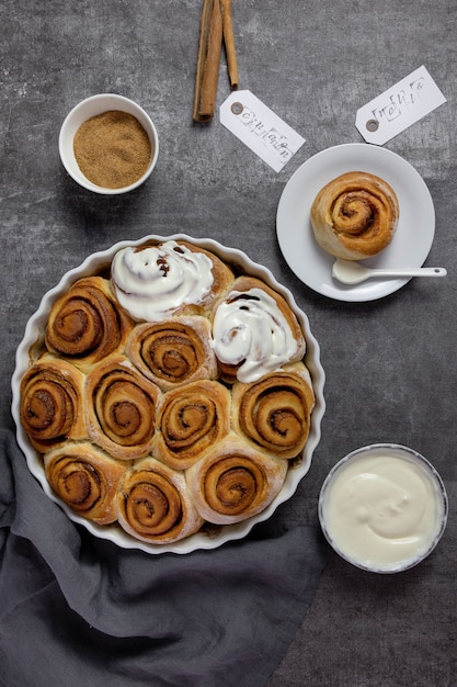 Zimtschnecken, Brötchen in einer Auflaufform mit braunem Zucker, Quark-Sahne-Sauce und Zimtstangen auf dunklem Beton.