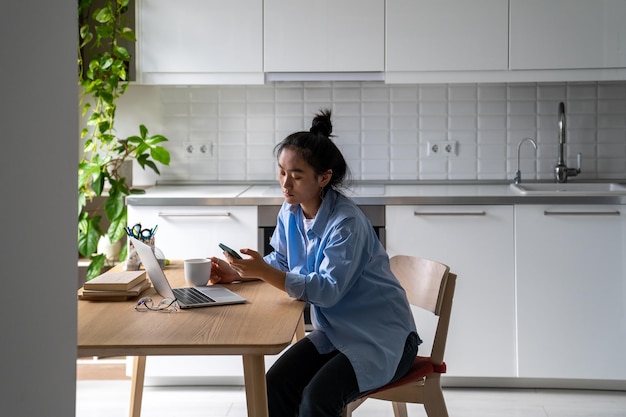 Zögernde asiatische Frau, die Social-Media-Feeds am Telefon durchsucht, sitzt mit Laptop am Küchentisch
