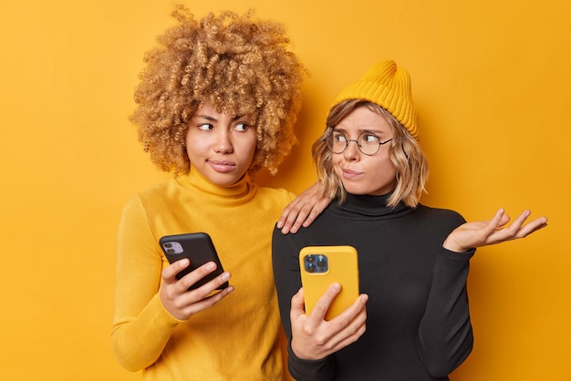 Foto zögernde junge zwei frauen schauen sich unsicher an, verwenden moderne mobiltelefone, verwenden neue smartphone-anwendung, tragen schwarz-gelben rollkragen-hut, pose im innenbereich, wissen nicht, wie man neue geräte verwendet