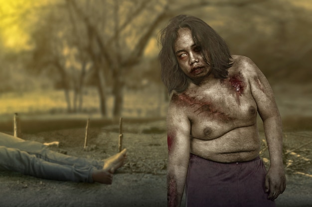 Zombie aterrador con sangre y herida en su cuerpo con un hombre muerto en el campo