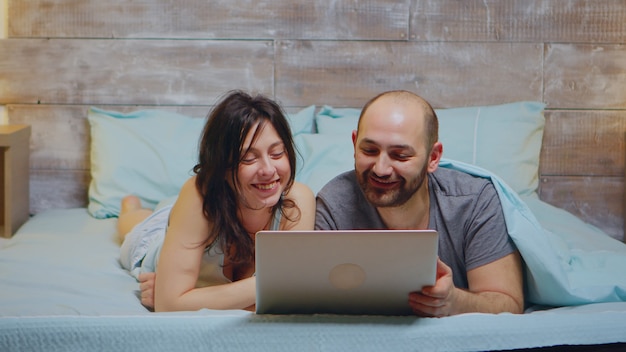 Zoom en la toma de una pareja en pijama riendo mientras usa la computadora portátil.