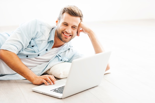 Zu Hause entspannen. Fröhlicher junger Mann, der am Laptop arbeitet und lächelt, während er in seiner Wohnung auf dem Boden liegt