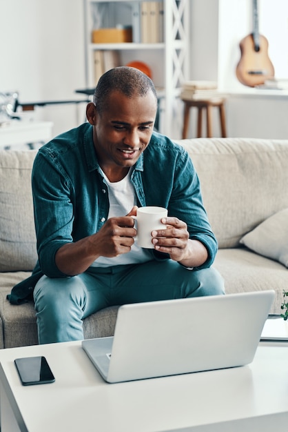 Zu Hause entspannen. Hübscher junger afrikanischer Mann, der Laptop benutzt und lächelt, während er drinnen sitzt