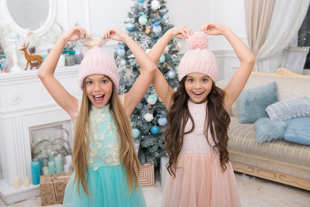 Zubehör für die Wintersaison. Kinder tragen Strickmützen. Mädchen lange Haare glücklich lächelnde Gesichter Weihnachtsbaum Hintergrund. Kinder tragen warme, weiche rosa Strickmützen. Ausgefallene Strickmützen für Mädchen. Trendige Mädchen.