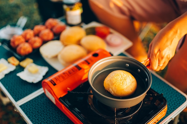 Zubereitung von Camping-Lebensmitteln. Burger auf Pfanne auf touristischem Feuerofen. Campkochen am Ufer des Sees.