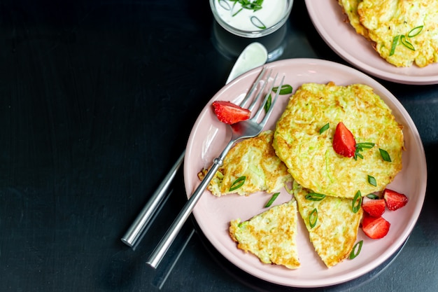 Zucchini-Krapfen, vegetarische Zucchini-Krapfen, serviert mit frischen Kräutern und saurer Sahne. Mit Erdbeeren und Frühlingszwiebeln garniert. Auf einem dunklen Tisch.