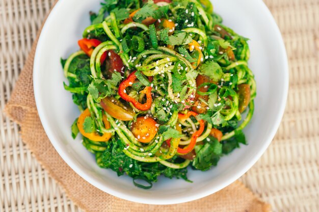 Zucchinispaghettisalat mit gelben Tomaten, rotem Pfeffer, Zwiebel, seasame Samen, Koriander in der weißen Schüssel. Frischer Salat auf dem Tisch. Gemüsegericht. Horizontaler Draufsichtschuß