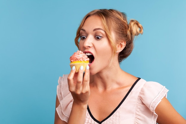 Zuckersucht. Porträt einer hungrigen blonden jungen erwachsenen Frau im Kleid, die köstlichen Kuchen beißt und mit dem Wunsch aussieht, süßes Dessert zu essen. Studioaufnahme im Innenbereich isoliert auf blauem Hintergrund.