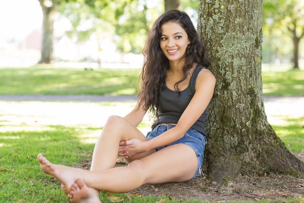 Zufälliger glücklicher Brunette, der das Lehnen am Baum sitzt