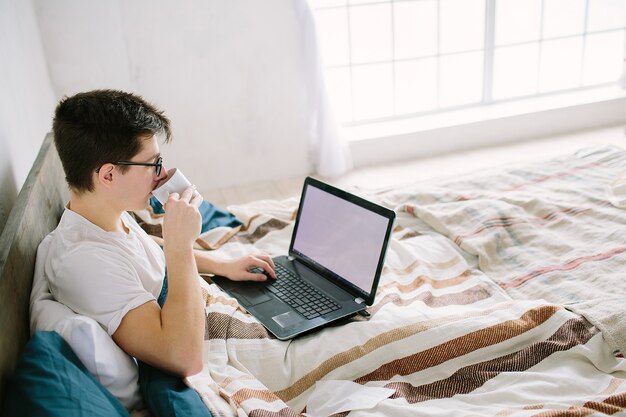 Zufälliger junger Mann, der Laptop im Bett zu Hause benutzt und Kaffee trinkt