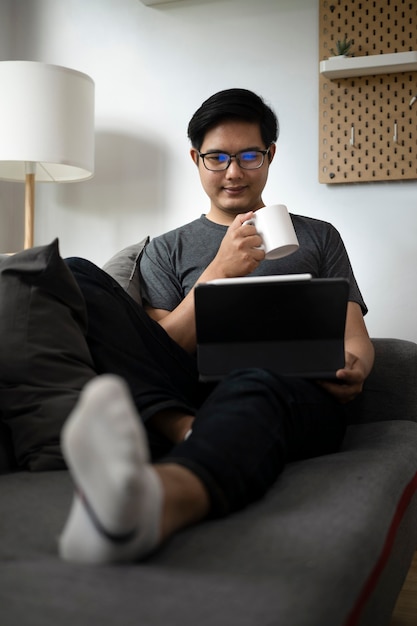 Zufälliger Mann, der Kaffeetasse hält und Computertablette auf Couch verwendet.