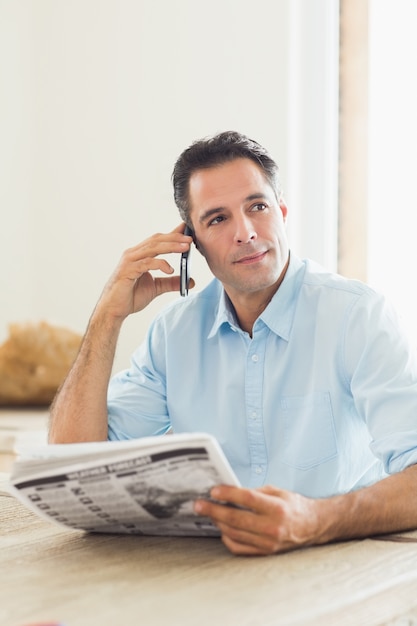 Zufälliger Mann mit der Zeitung, die Mobiltelefon in der Küche verwendet