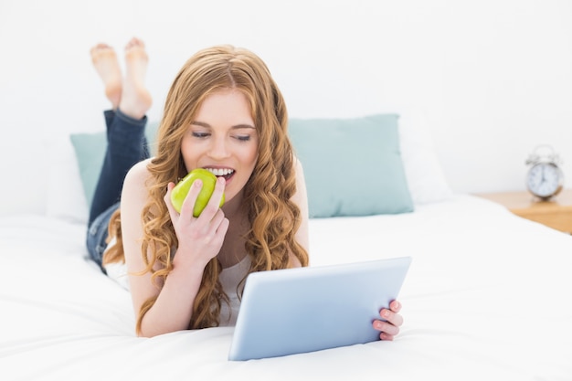 Zufälliges blondes unter Verwendung des Tablet-PCs beim Essen des Apfels im Bett