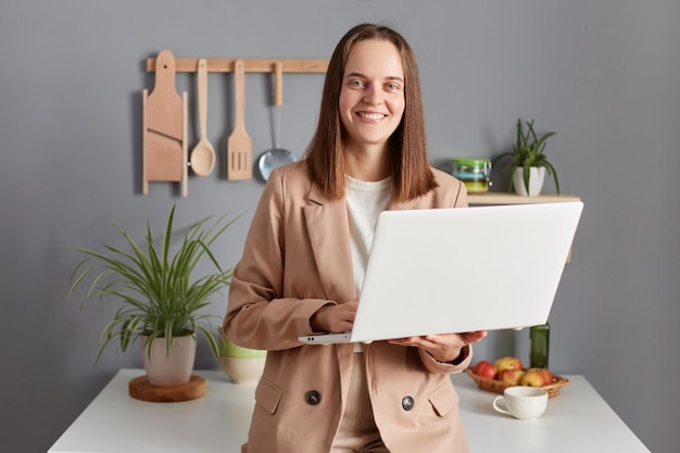 Zufrieden lächelnde Freiberuflerin mit braunen Haaren in beiger Jacke, die im Kücheninnenraum steht und von zu Hause aus am Laptop arbeitet und sich auf ein Online-Meeting vorbereitet