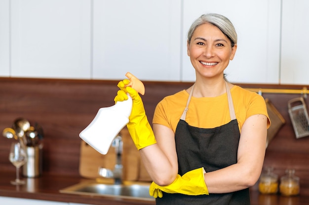 Foto zufrieden lächelnde reife asiatische hausfrau oder reinigungsdame in gelben handschuhen und schürze mit reinigungsmittel bereit zu hause oder im büro reinigung schaut in die kamera