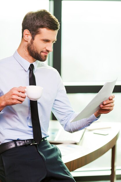 Zufrieden mit der geleisteten Arbeit. Hübscher junger Mann in Hemd und Krawatte, der Kaffeetasse hält und Dokument untersucht, während er sich am Schreibtisch lehnt