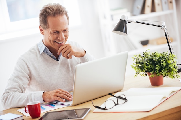 Zufrieden mit seiner Arbeit. Fröhlicher reifer Mann, der am Laptop arbeitet und lächelt, während er an seinem Arbeitsplatz sitzt