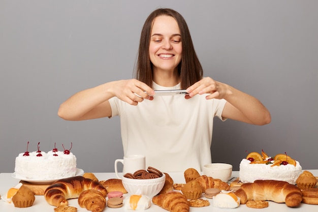 Zufriedene Bloggerin im weißen T-Shirt fotografiert zuckerhaltige Produkte und erstellt Inhalte für ihren kulinarischen Vlog isoliert vor grauem Hintergrund