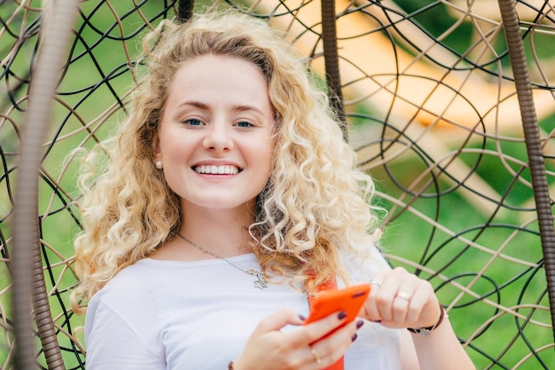 Zufriedene blonde junge frau mit lockigem haar hat ein positives lächeln nutzt ein modernes handy zum chatten mit freunden kann sich ihr leben ohne moderne technologie nicht vorstellen sieht positiv in die kamera