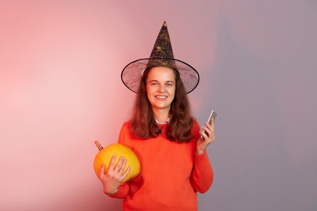 Zufriedene Frau in Hexenhut mit orangefarbenem Kürbis und Smartphone in den Händen, die mit einem Lächeln in die Kamera blickt und in festlicher Halloween-Stimmung isoliert auf Neonlichthintergrund posiert
