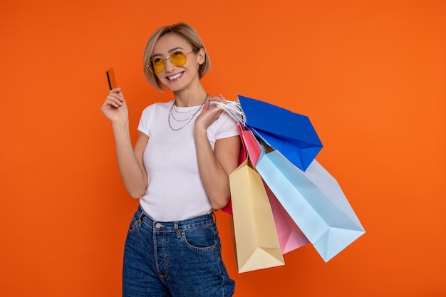 Zufriedene Frau in weißem T-Shirt und Jeans mit Einkaufstaschen und Kreditkarte, isoliert über orangefarbenem Hintergrund