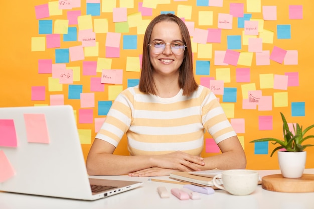 Zufriedene, freundliche Managerin in gestreiftem T-Shirt, die am Tisch im Büro vor einem Laptop an der gelben Wand sitzt, mit Aufklebern, die mit einem zahnigen Lächeln in die Kamera schauen und bereit sind, ihre Arbeit zu beginnen