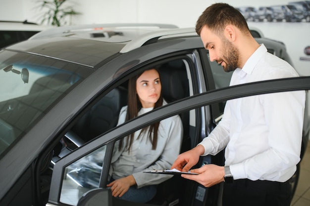 Zufriedene, glückliche kaukasische weibliche Kundin Kundenfrau sitzt am Lenkrad eines neuen Autos, während der männliche Verkäufer ihr hilft, es auszuwählen
