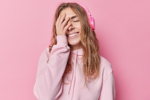 Zufriedene junge Frau bedeckt das Gesicht mit der Hand, lächelt breit, schließt die Augen, hat eine optimistische Stimmung, gekleidet in einen lässigen Hoodie, hört Audiospur über Kopfhörer, die über rosafarbenem Hintergrund isoliert sind. Emotionen-Konzept