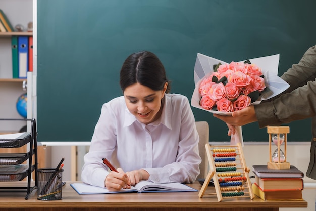 Zufriedene junge Lehrerin, die auf ein Notizbuch schreibt, erhielt einen Blumenstrauß, der mit Schulwerkzeugen im Klassenzimmer am Schreibtisch saß