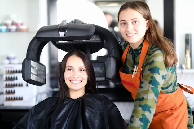 Zufriedene Kunden sitzen auf einem Stuhl in Umhang, trockenem Haar und schauen in der Nähe des Friseursalons weg.