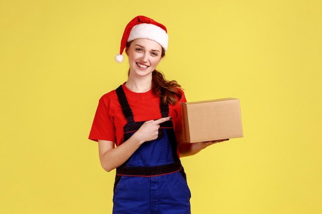 Zufriedene Lieferfrau, die auf das Paket zeigt, schnelle Lieferung von Geschenken für den Winterurlaub, blaue Overalls und Weihnachtsmannmütze trägt. Studioaufnahme im Innenbereich isoliert auf gelbem Hintergrund.
