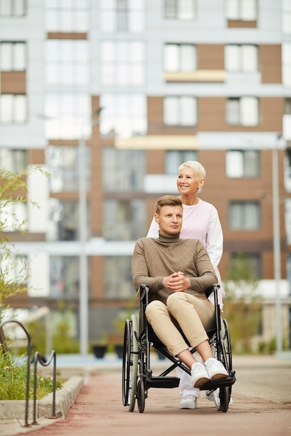 Zufriedene reife professionelle Krankenschwester mit blonden Haaren, die Rollstuhl mit unzufriedenem Patienten schiebt, während t