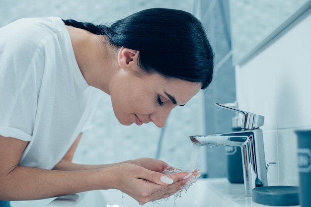 Zufriedene schöne Frau, die ihre Augen schließt und lächelt, während sie mit ihren Händen unter fließendem Wasser vor einem Waschbecken steht