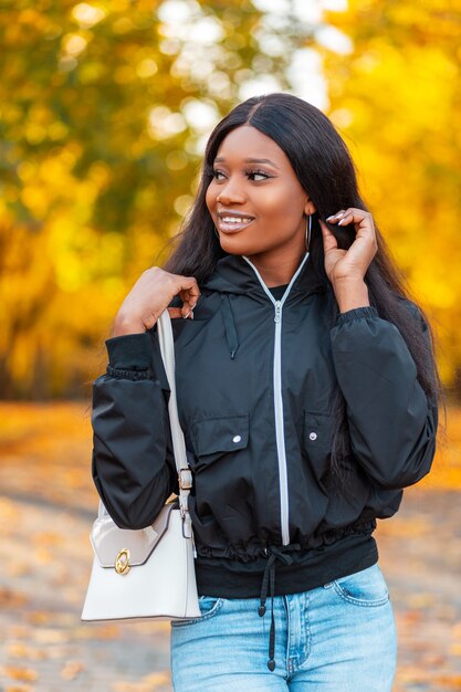 Zufriedene schöne junge lächelnde schwarze Frau in einer modischen Jacke mit Blue Jeans und einer stilvollen Handtasche spaziert in der Natur vor einem Hintergrund von leuchtend gelbem Herbstlaub