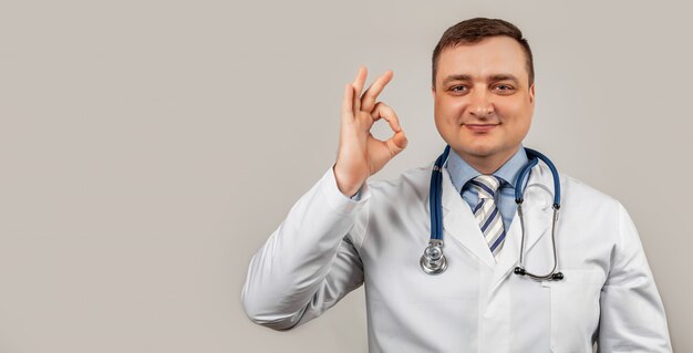 Zufriedener Arzt Mann in einem medizinischen Mantel und mit einem Stethoskop zeigt okay okay Zeichen der Zustimmung lächelt, weißer Hintergrund