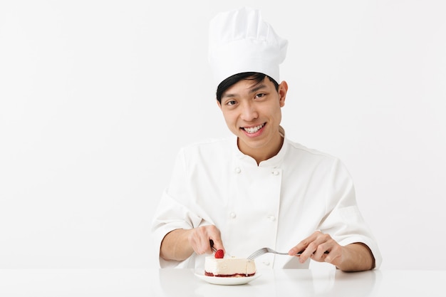 zufriedener asiatischer Hauptmann in der weißen Kochuniform, die in die Kamera lächelt, während sie leckeren Käsekuchen isst, der über weißer Wand lokalisiert wird