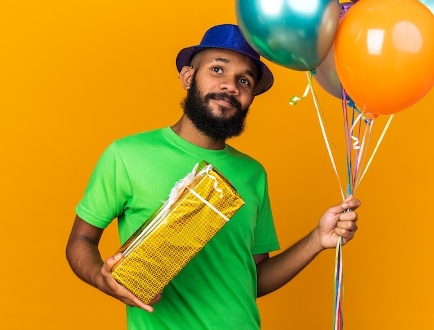 Zufriedener junger afroamerikaner mit partyhut, der luftballons mit geschenkbox isoliert auf oranger wand hält