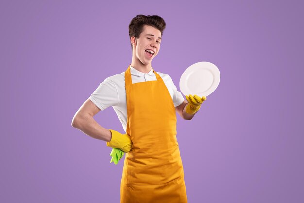 Zufriedener junger Hausmann in Schürze mit Hand auf Hüfte und Teller, Blick in die Kamera auf violettem Hintergrund