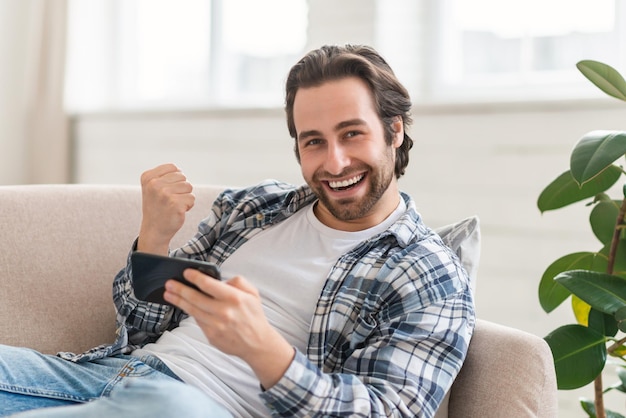 Zufriedener junger Kaukasier auf dem Sofa mit Smartphone, der mit der Hand ein Siegeszeichen macht, genießt den Erfolg beim Sieg